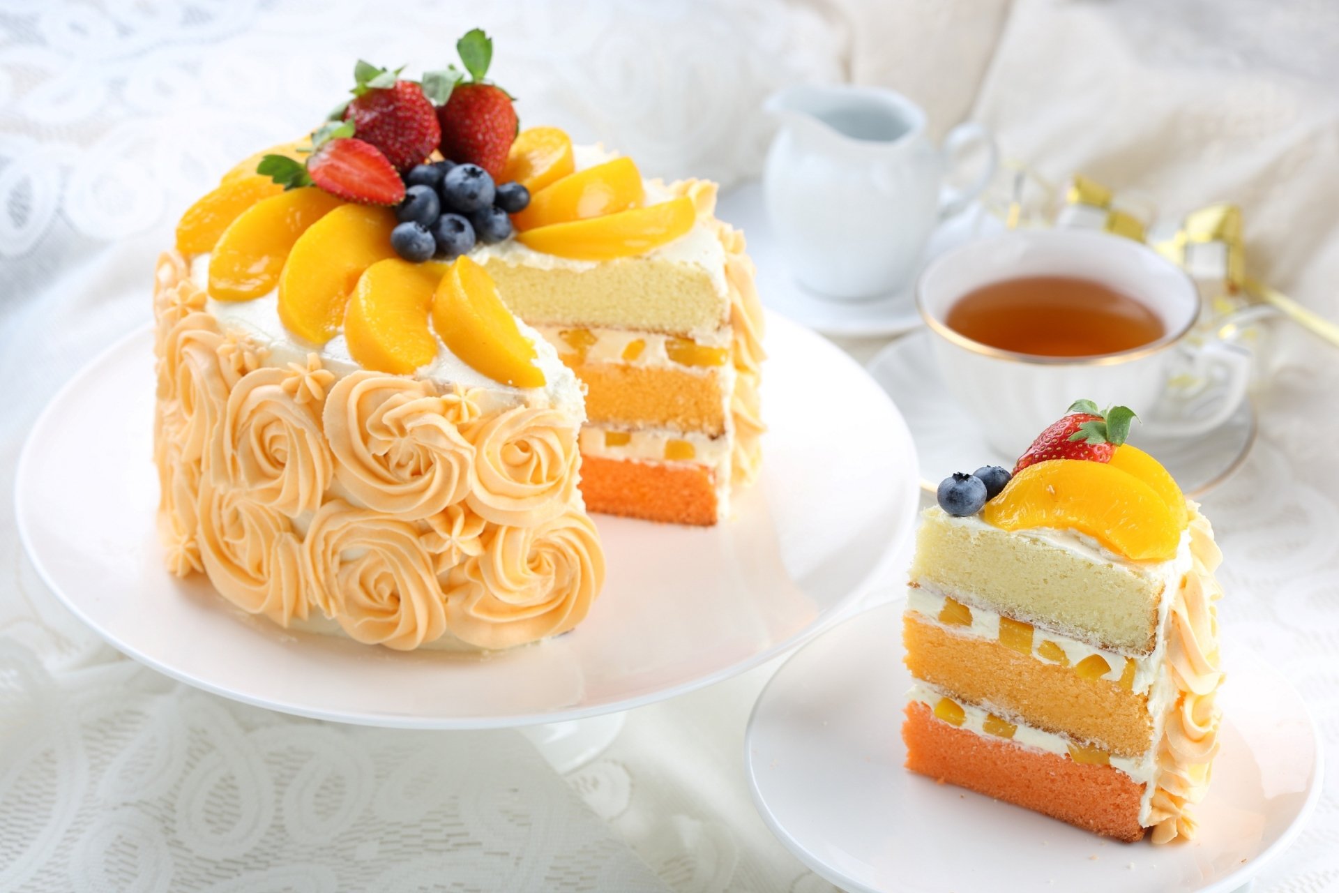 HD desktop wallpaper featuring a beautifully decorated fruit cake with peaches, strawberries, and blueberries, accompanied by a cup of tea in a bright still life setting.