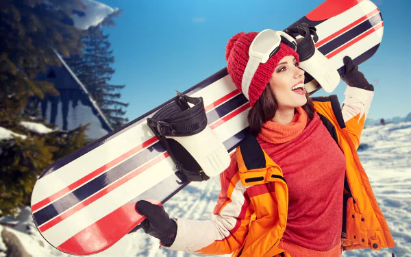 Smiling snowboarder in a red hat and orange jacket carries a snowboard on a sunny winter day, captured in 4K Ultra HD for a vibrant sports desktop background.