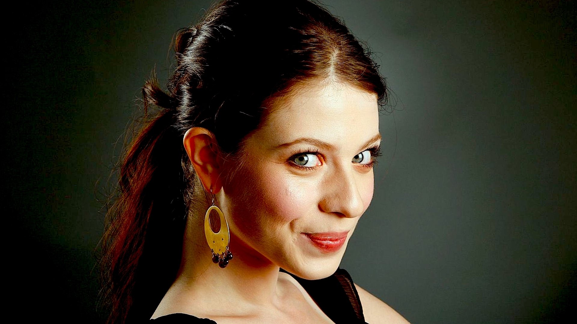 HD desktop wallpaper of a smiling young woman with dark hair pulled back, wearing gold oval earrings and a black top, looking over her shoulder against a gray background.