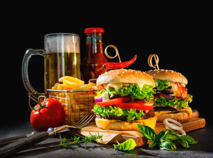 A vibrant still life of two gourmet burgers with lettuce, tomato, and onions, accompanied by a basket of French fries, a glass of beer, and a bottle of ketchup on a dark background.