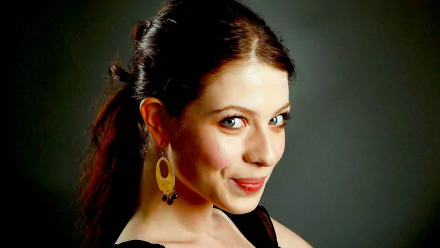 HD desktop wallpaper of a smiling young woman with dark hair pulled back, wearing gold oval earrings and a black top, looking over her shoulder against a gray background.