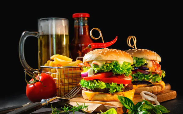 A vibrant still life of two gourmet burgers with lettuce, tomato, and onions, accompanied by a basket of French fries, a glass of beer, and a bottle of ketchup on a dark background.
