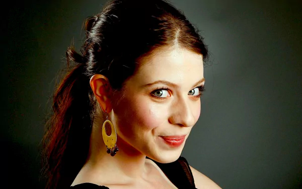 HD desktop wallpaper of a smiling young woman with dark hair pulled back, wearing gold oval earrings and a black top, looking over her shoulder against a gray background.