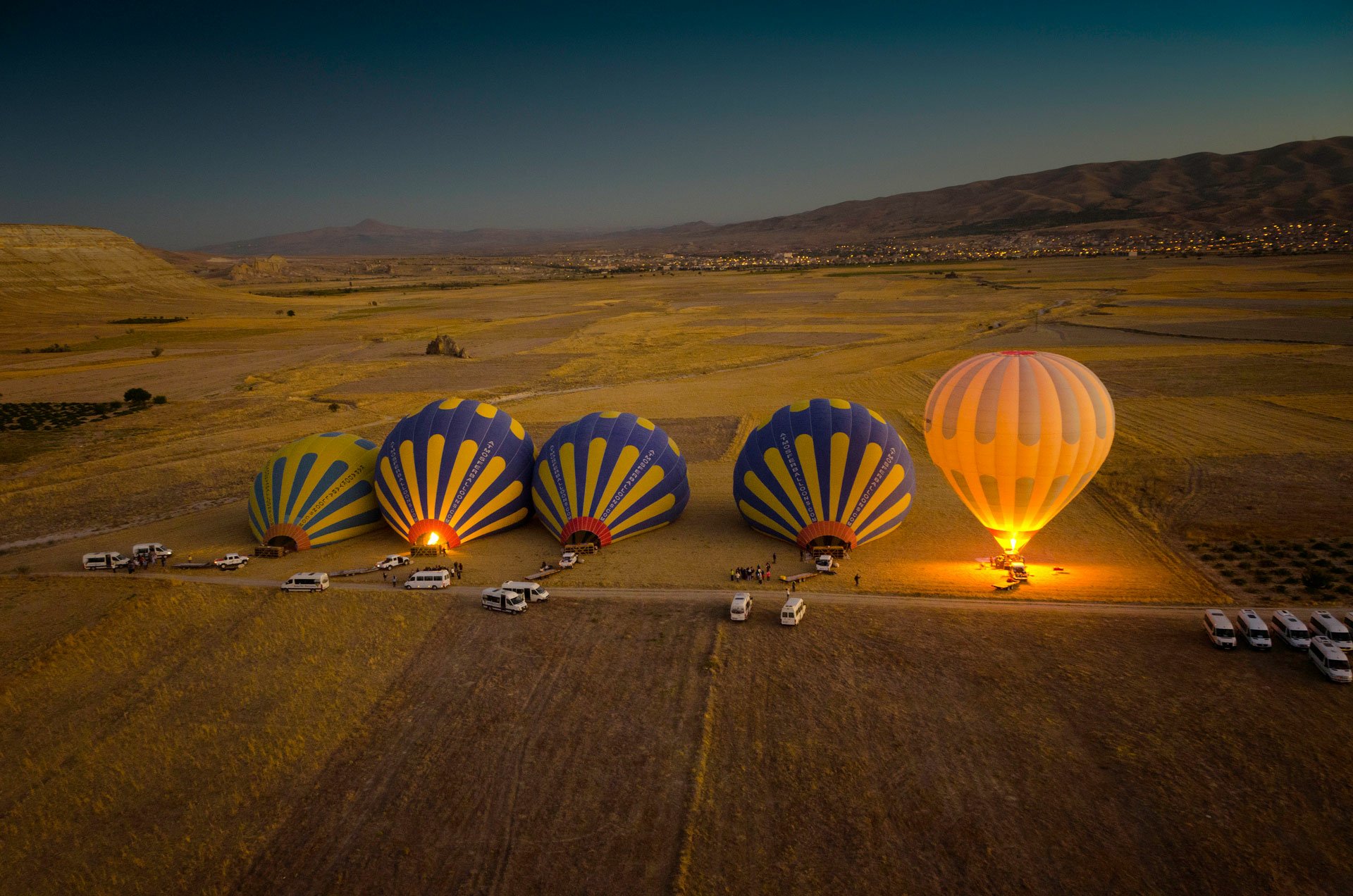 A tilt-shift HD desktop wallpaper featuring several hot air balloons being prepared for flight beside vehicles in a vast, open landscape at dusk.