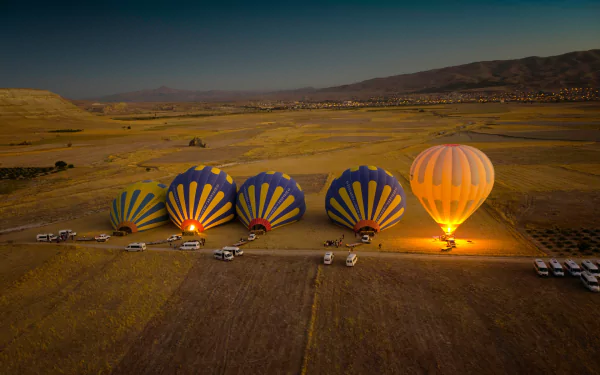 A tilt-shift HD desktop wallpaper featuring several hot air balloons being prepared for flight beside vehicles in a vast, open landscape at dusk.