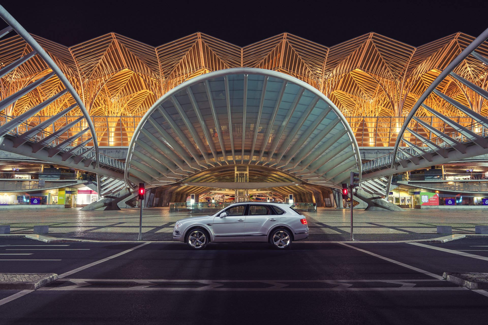 Silver Bentley Bentayga SUV centered in front of illuminated contemporary architectural terminal at night — 4K Ultra HD PC desktop wallpaper background.