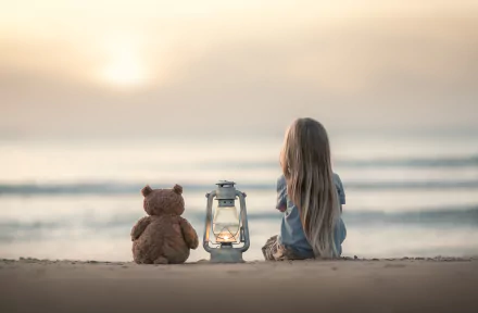 A little blonde girl with long hair sits on the beach at sunset, accompanied by a teddy bear and a lantern, captured with depth of field in this HD desktop wallpaper.