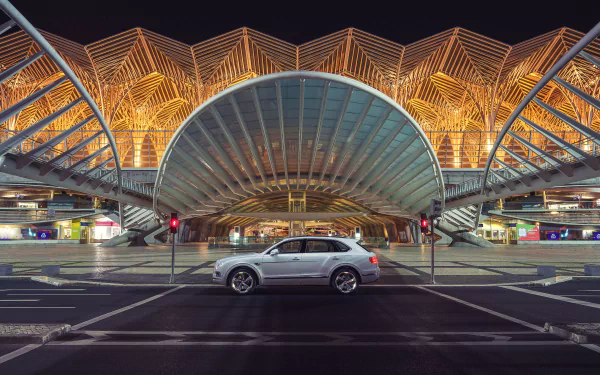Silver Bentley Bentayga SUV centered in front of illuminated contemporary architectural terminal at night — 4K Ultra HD PC desktop wallpaper background.
