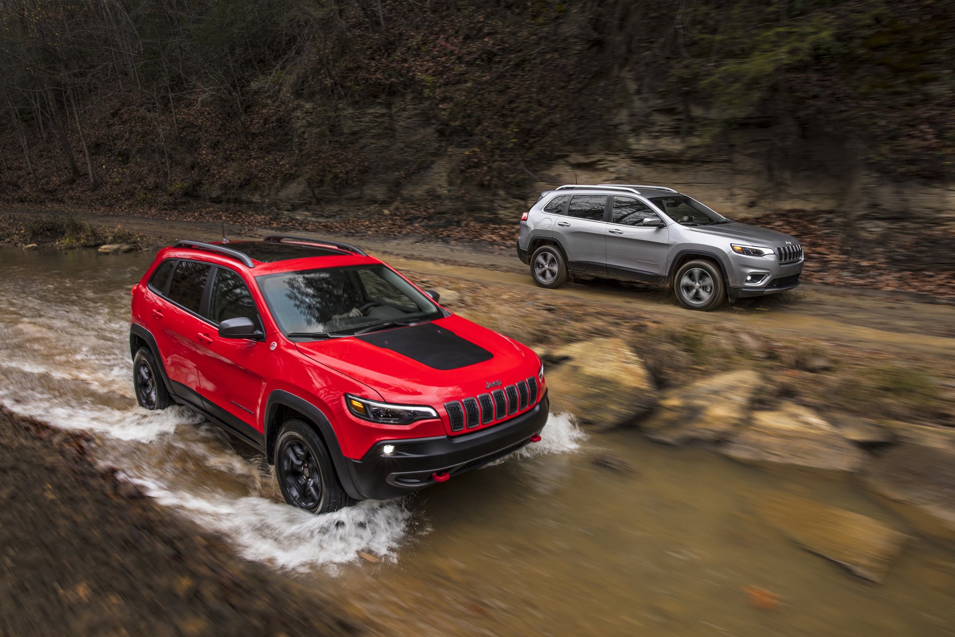 Two Jeep Cherokee SUVs — a silver car and a red Jeep — splashing through a rocky stream; 2K Quad HD PC desktop wallpaper/background.