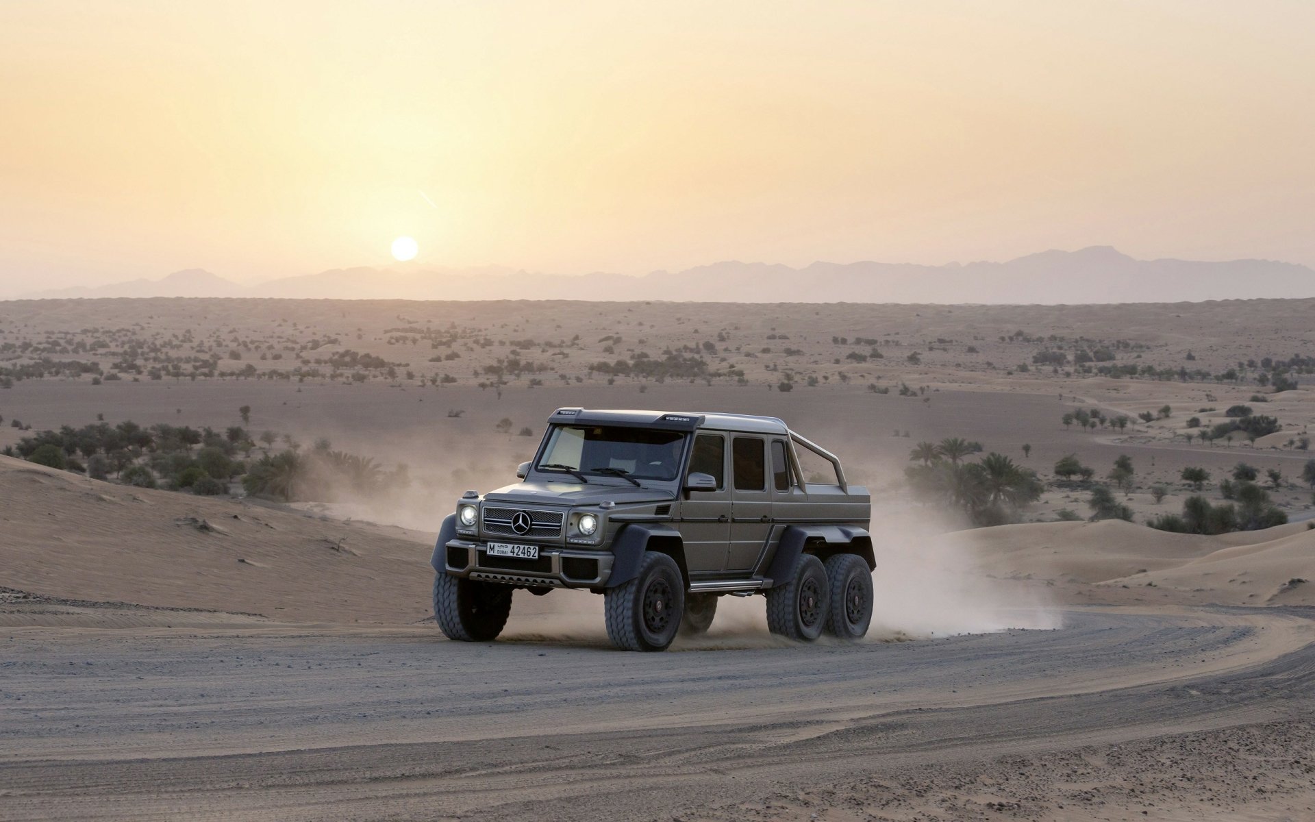A Mercedes-Benz G-Class vehicle drives through a desert landscape at sunset, kicking up dust with mountains in the background.