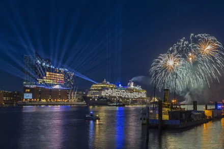 Night view of Hamburg, Germany featuring illuminated buildings, a docked cruise ship, vehicles by the water, and vibrant fireworks lighting up the sky.
