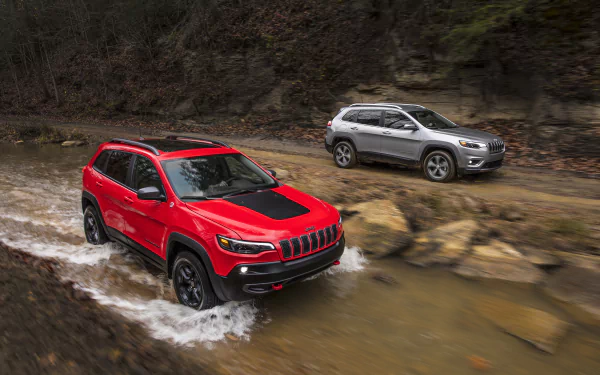 Two Jeep Cherokee SUVs — a silver car and a red Jeep — splashing through a rocky stream; 2K Quad HD PC desktop wallpaper/background.