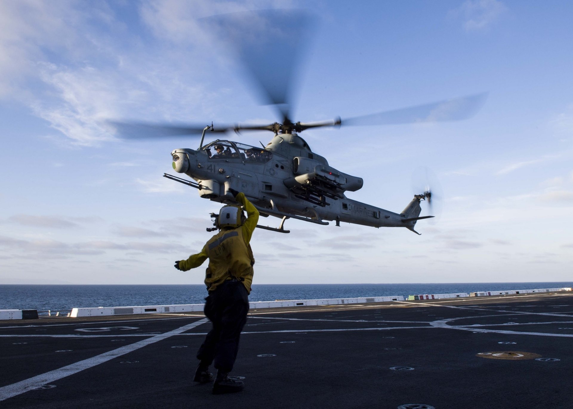 Bell AH-1 Cobra attack helicopter hovering over an aircraft carrier deck with a deck crew member, military aircraft in HD desktop wallpaper