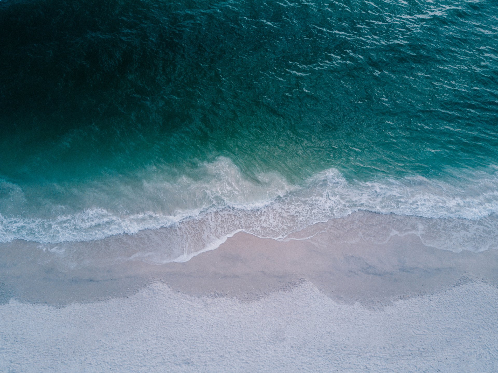 Aerial view of turquoise ocean waves gently rolling onto a sandy beach, captured in stunning 4K Ultra HD for a vibrant PC desktop wallpaper.