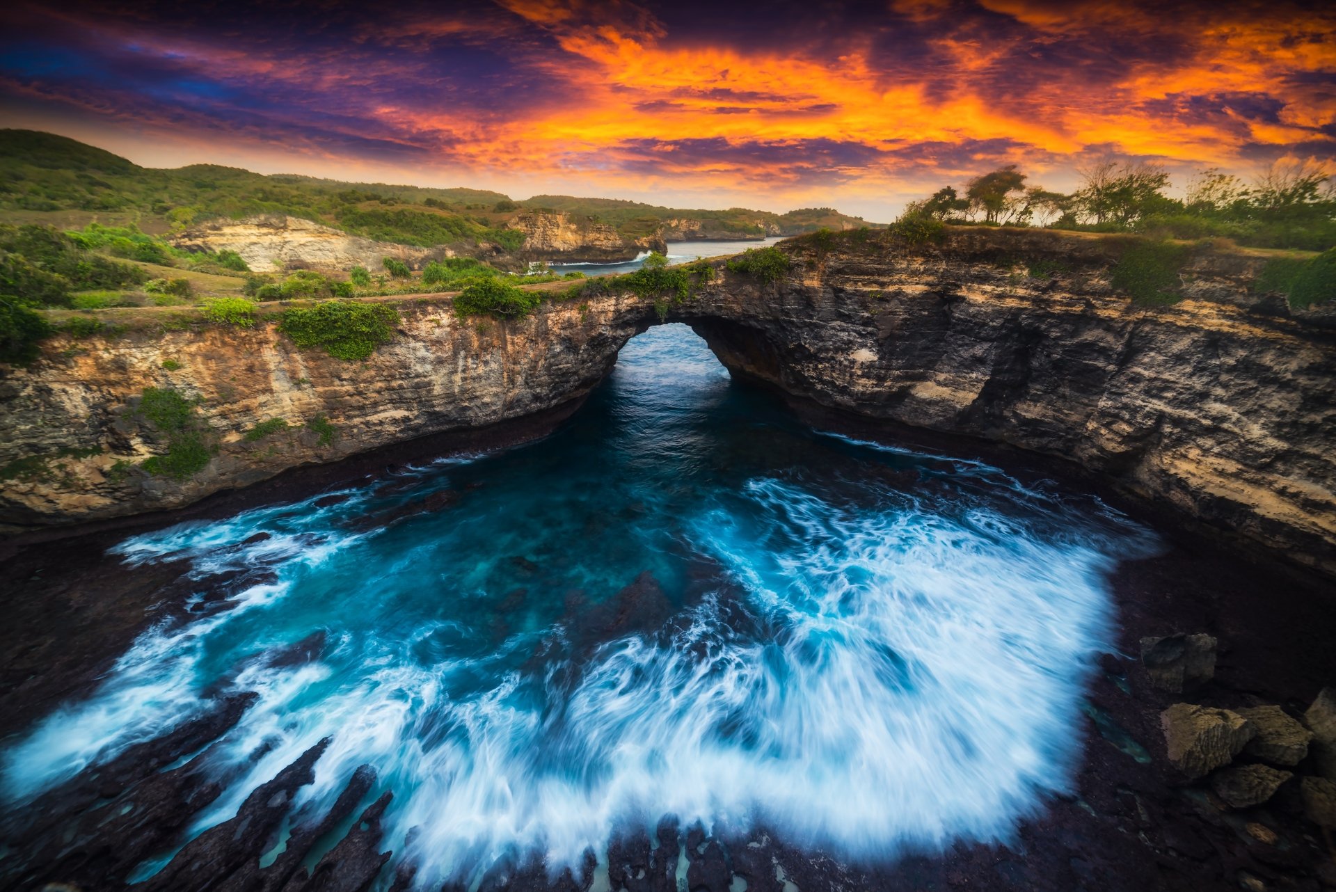 4K Ultra HD desktop wallpaper of a natural coastal arch with waves crashing beneath a vibrant sunset sky over rugged cliffs and lush greenery.