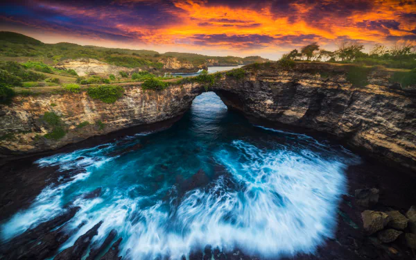 4K Ultra HD desktop wallpaper of a natural coastal arch with waves crashing beneath a vibrant sunset sky over rugged cliffs and lush greenery.