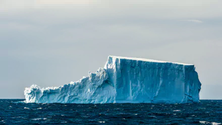 4K Ultra HD desktop wallpaper of a massive iceberg floating in the dark blue ocean under a cloudy sky, showcasing the stark beauty of nature.