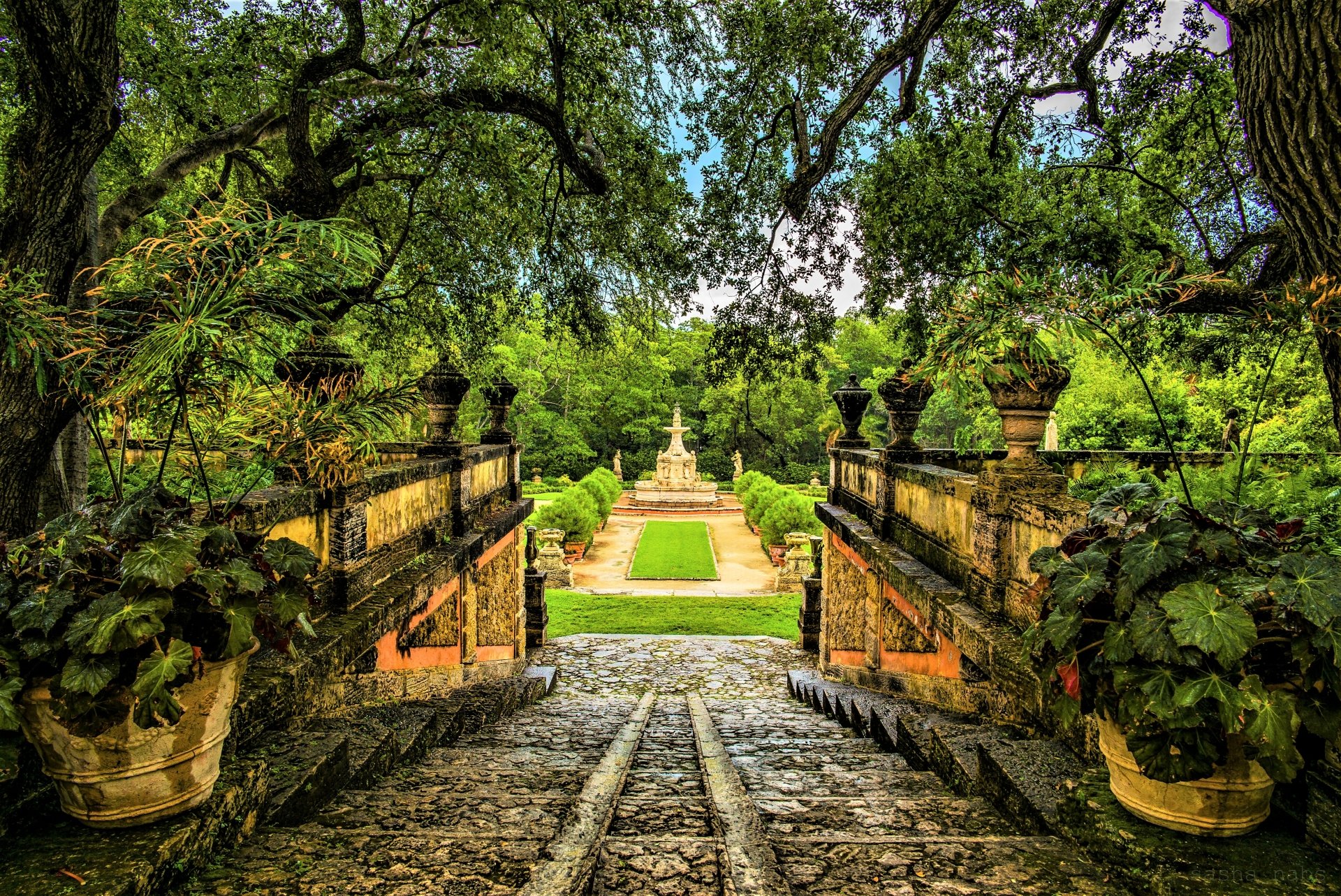 A lush green park in Florida featuring a cobblestone pathway framed by large trees and historic stone railings, captured in vibrant 4K Ultra HD detail.
