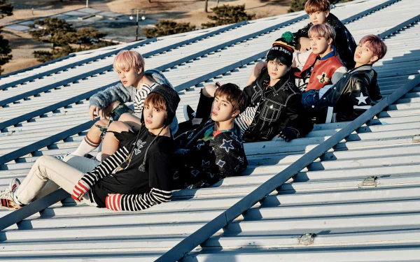 A high-definition desktop wallpaper featuring the music group BTS, sitting on a slanted rooftop with trees and a clear sky in the background.