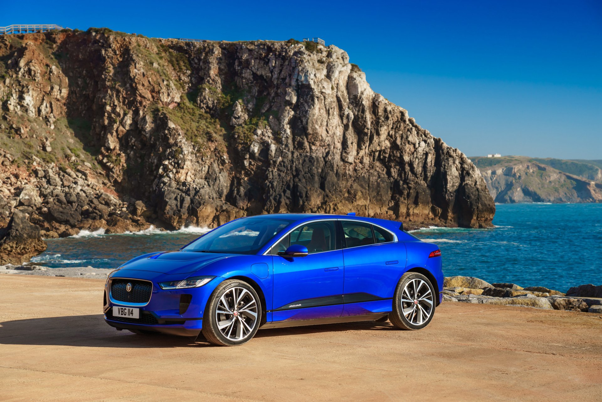 Blue Jaguar I-Pace vehicle parked on a coastal road with rocky cliffs and ocean in the background, captured in vibrant 4K Ultra HD detail.