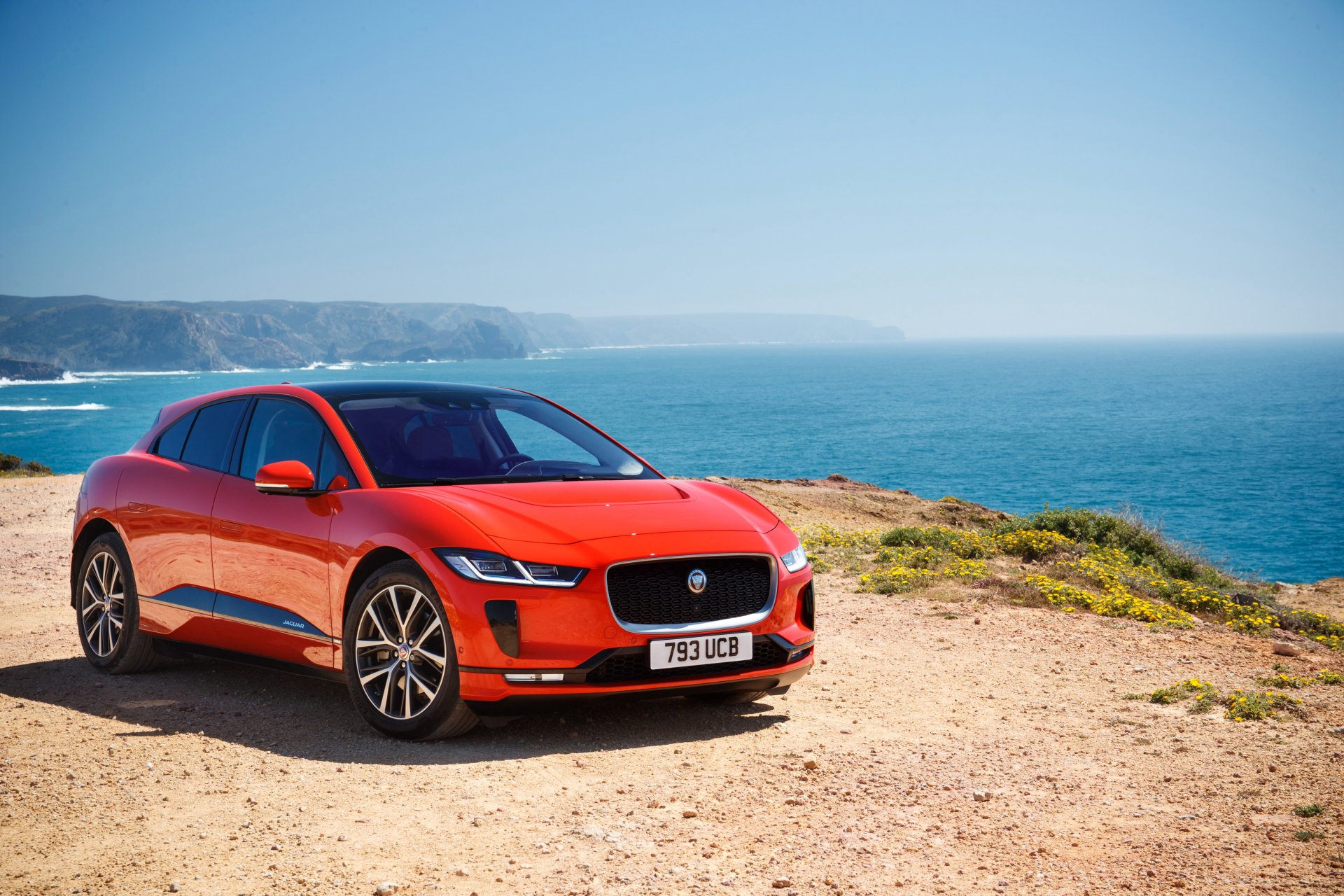 A red Jaguar I-Pace electric vehicle parked on a coastal cliff with the ocean and distant hills in the background, shown in 4K Ultra HD.