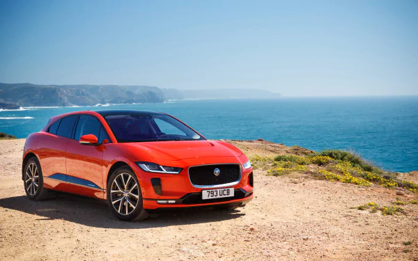A red Jaguar I-Pace electric vehicle parked on a coastal cliff with the ocean and distant hills in the background, shown in 4K Ultra HD.