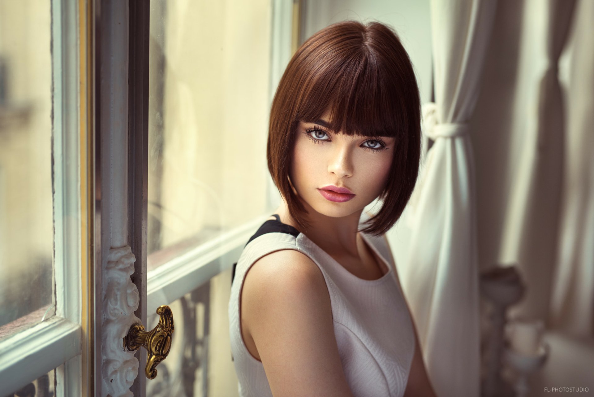 HD desktop wallpaper of Marie Grippon, a French model with short brunette hair, blue eyes, and wearing lipstick, posing indoors near a window with elegant curtains.