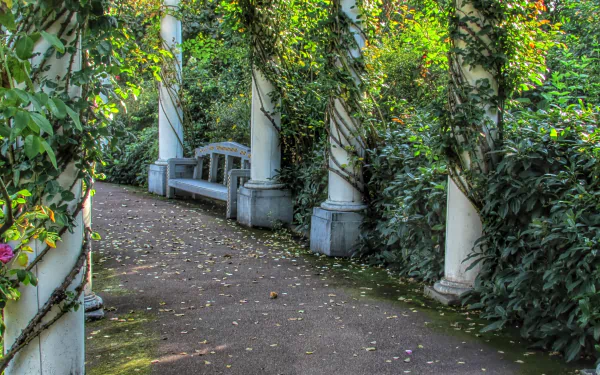 A serene man-made pathway in spring, lined with stone columns wrapped in vines and surrounded by lush greenery and blooming flowers. HD PC desktop wallpaper.