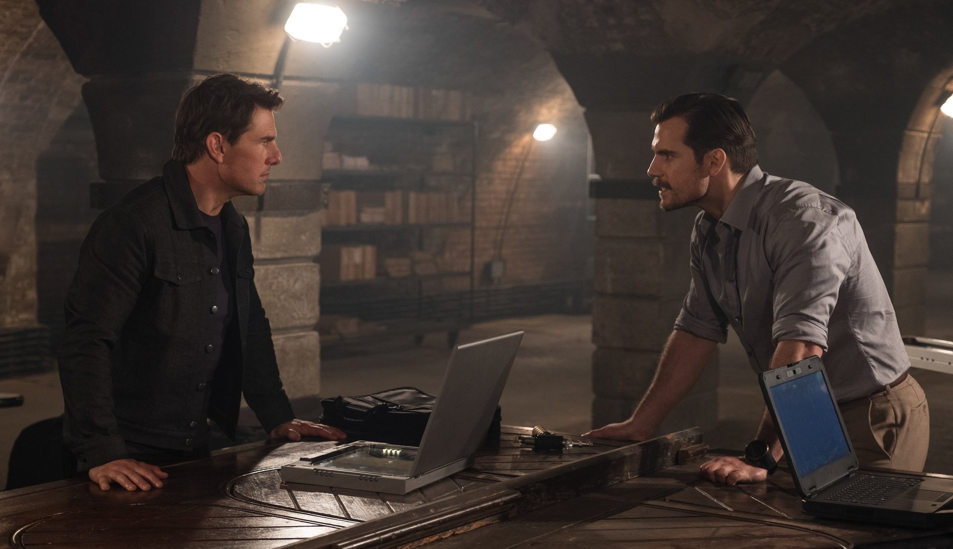 Mission: Impossible - Fallout still: two men lean across a wooden table in a dim basement, laptops open in a tense standoff — 5K Ultra HD PC desktop wallpaper.