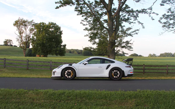 White Porsche 911 GT3 vehicle parked on a country road beside a wooden fence and green fields under a blue sky — 5K Ultra HD PC desktop wallpaper/background.