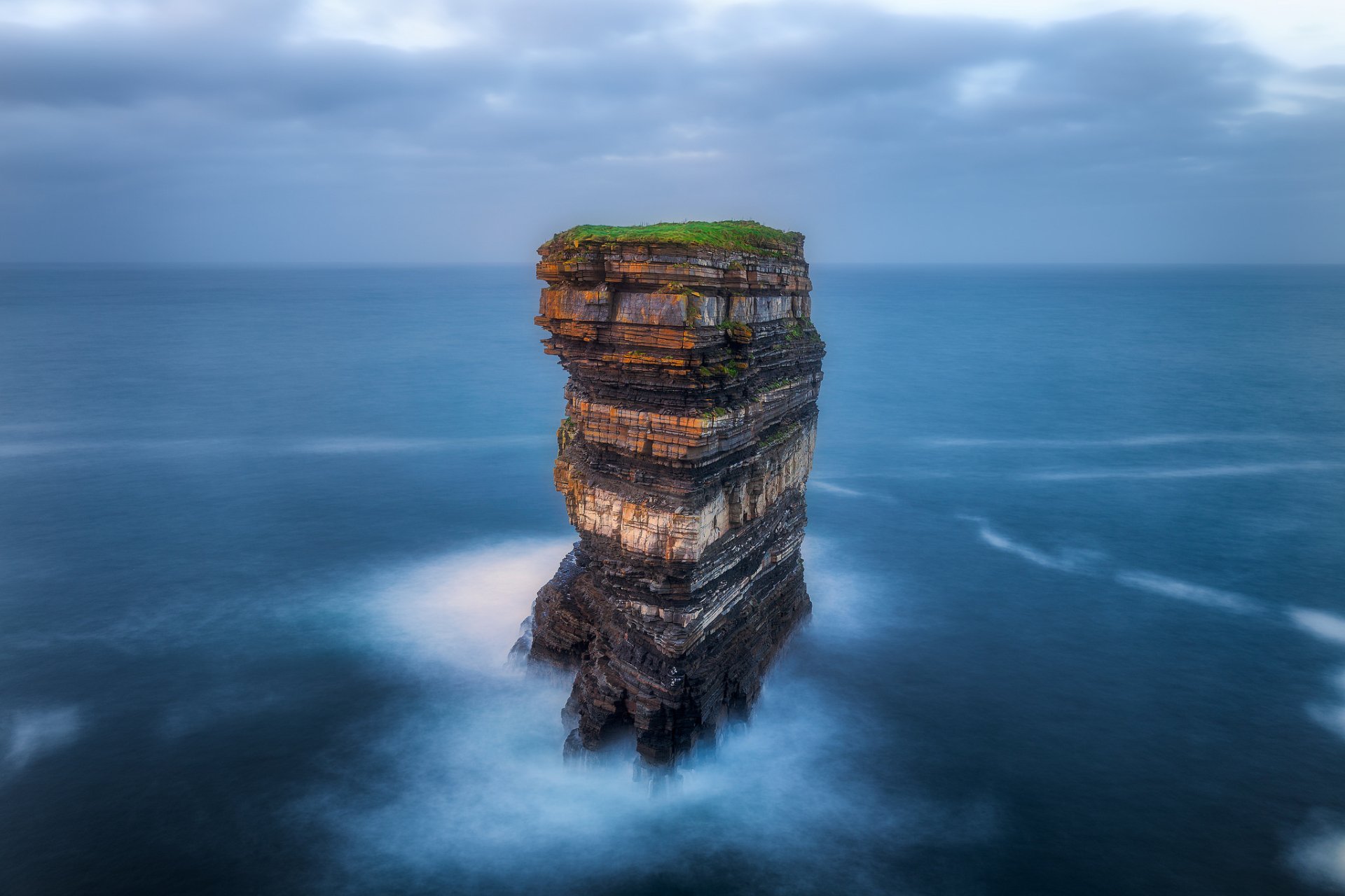 HD desktop wallpaper showcasing a solitary rock formation rising from the ocean near Ireland, with a misty sea and distant horizon under a cloudy sky.
