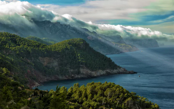 HD desktop wallpaper showcasing the stunning coastline of Mallorca, Spain, with lush forests and mountains draped in clouds overlooking the serene sea.