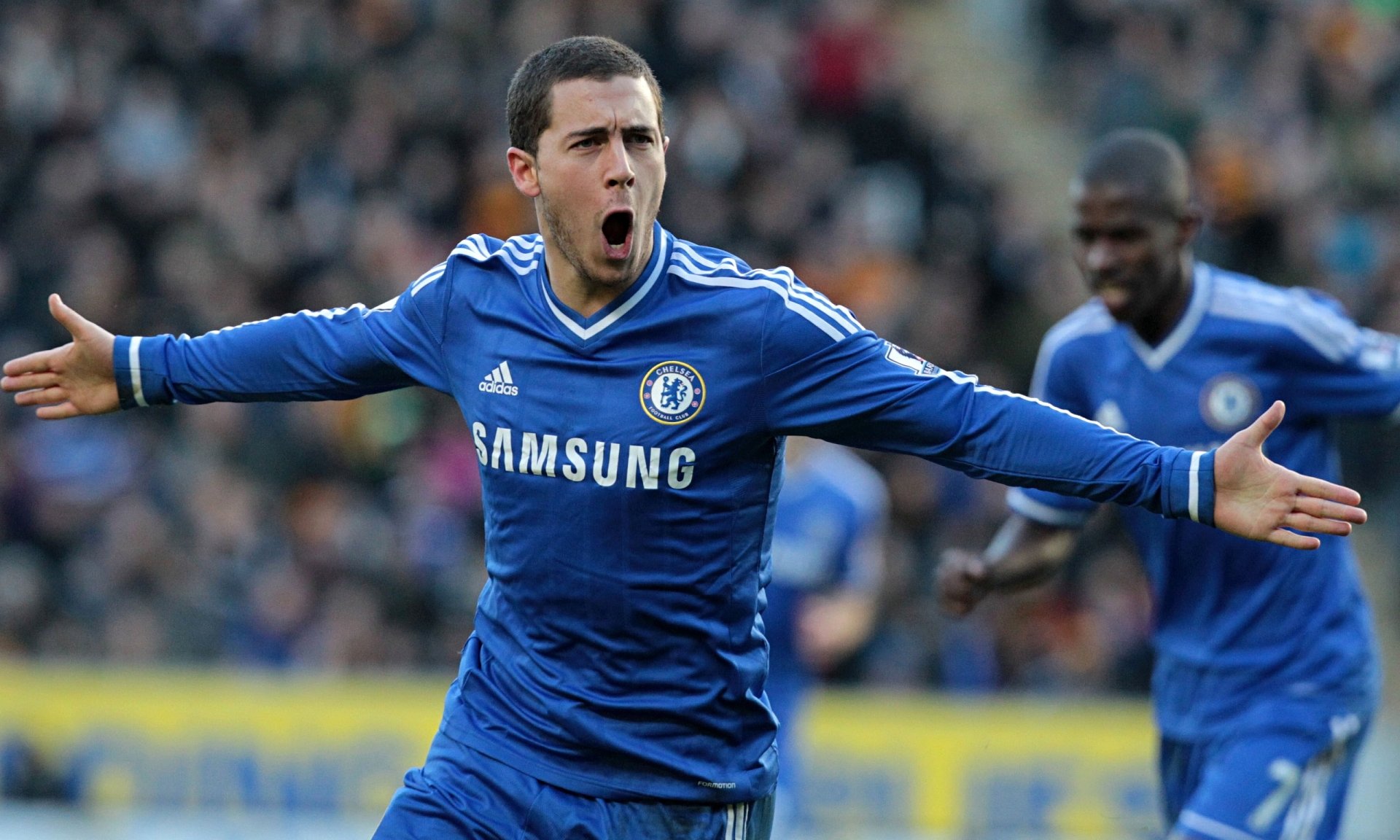 HD desktop wallpaper featuring Eden Hazard in a blue Chelsea kit, arms spread wide in celebration on the pitch.