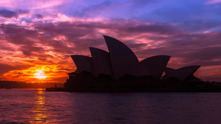 HD wallpaper of the Sydney Opera House silhouetted against a vibrant sunset sky, with clouds reflecting over calm waters in Sydney, Australia.