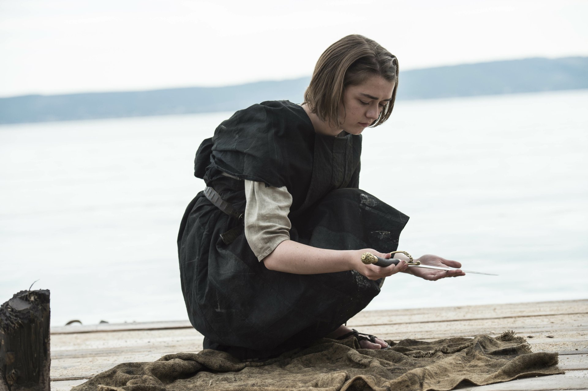 Arya Stark from the Game of Thrones TV show — 4K Ultra HD PC desktop wallpaper of her kneeling on a shore in dark clothes, examining small objects in her hands.