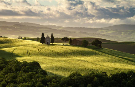 Sunlit rolling fields in Tuscany, Italy, with a small chapel and trees under a dramatic sky, captured in a stunning HD landscape photography wallpaper.