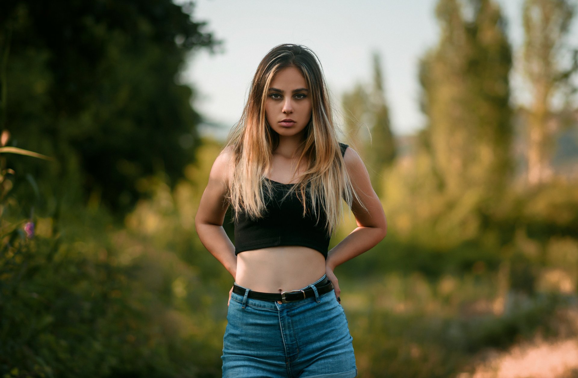 Blonde woman model stands confidently in a natural setting with blurred background, captured in HD with depth of field, designed as a PC desktop wallpaper.