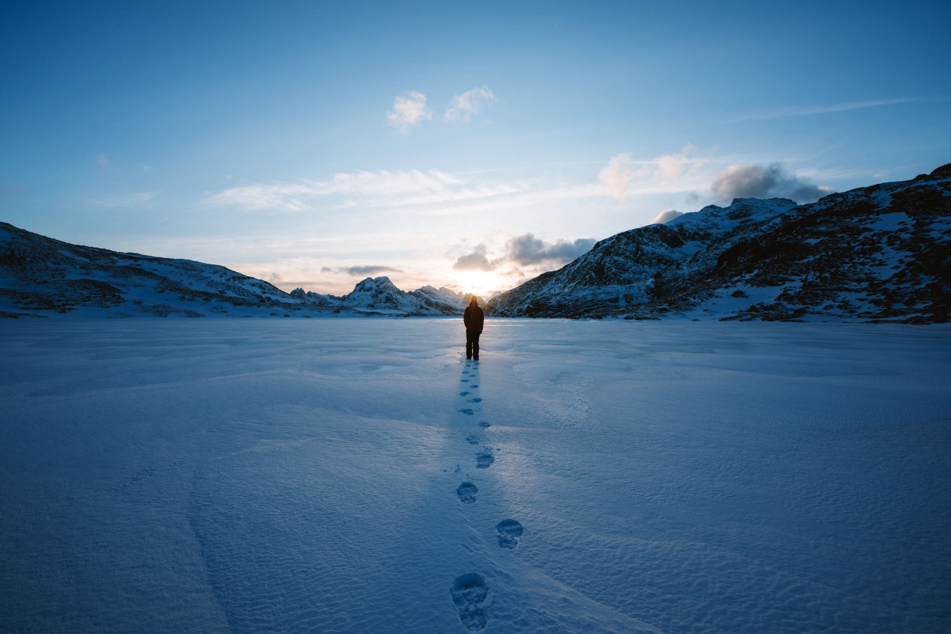 HD desktop wallpaper featuring a lone person standing on a vast snow-covered landscape with footprints leading back, framed by distant mountains under a clear sky.