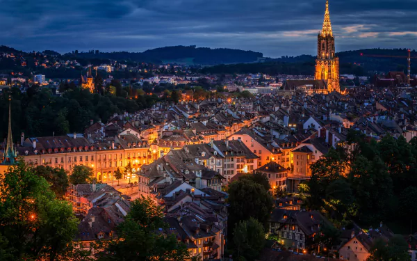 HD image of Bern, Switzerland at night, featuring a cityscape with illuminated buildings and a prominent lit tower. The background showcases hills under a dark, cloudy sky.