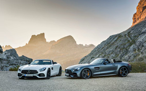 HD desktop wallpaper featuring a white and a silver Mercedes-AMG GT against a stunning mountainous backdrop at sunset.