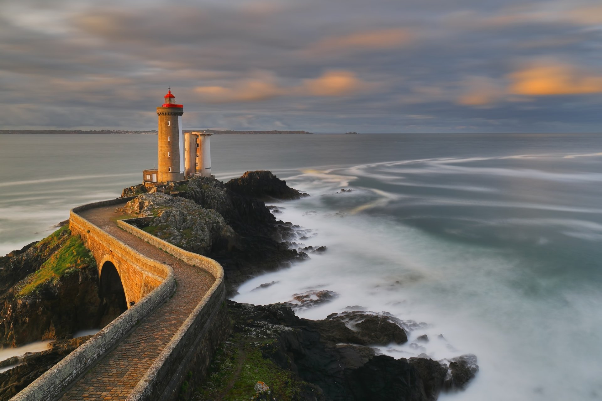 HD desktop wallpaper showing a lighthouse on a rocky path overlooking the ocean at the horizon, with a man-made building nearby. The sky is overcast, adding a dramatic touch to the scene.