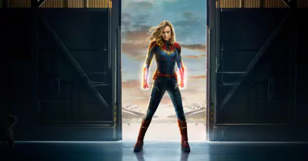 Brie Larson as Captain Marvel stands confidently in a dramatic pose, framed by open doors, showcasing her vibrant superhero suit against a stunning sky backdrop.