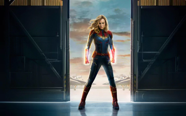 Brie Larson as Captain Marvel stands confidently in a dramatic pose, framed by open doors, showcasing her vibrant superhero suit against a stunning sky backdrop.