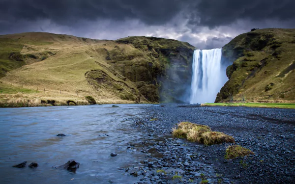 HD desktop wallpaper featuring Skógafoss waterfall in Iceland, with dramatic skies, rugged terrain, and a serene river flowing in the foreground.