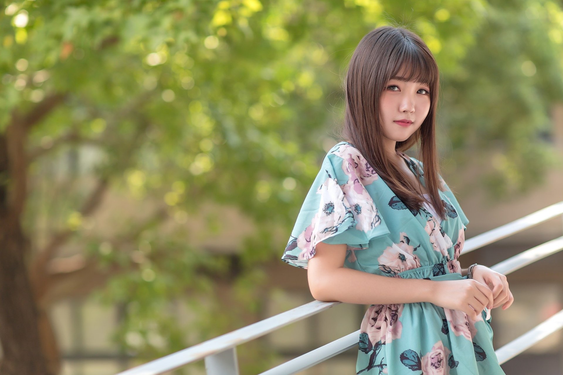 An Asian woman with a brunette hairstyle poses in a floral dress against a blurred green background. The depth of field highlights her as the focal point. HD desktop wallpaper.