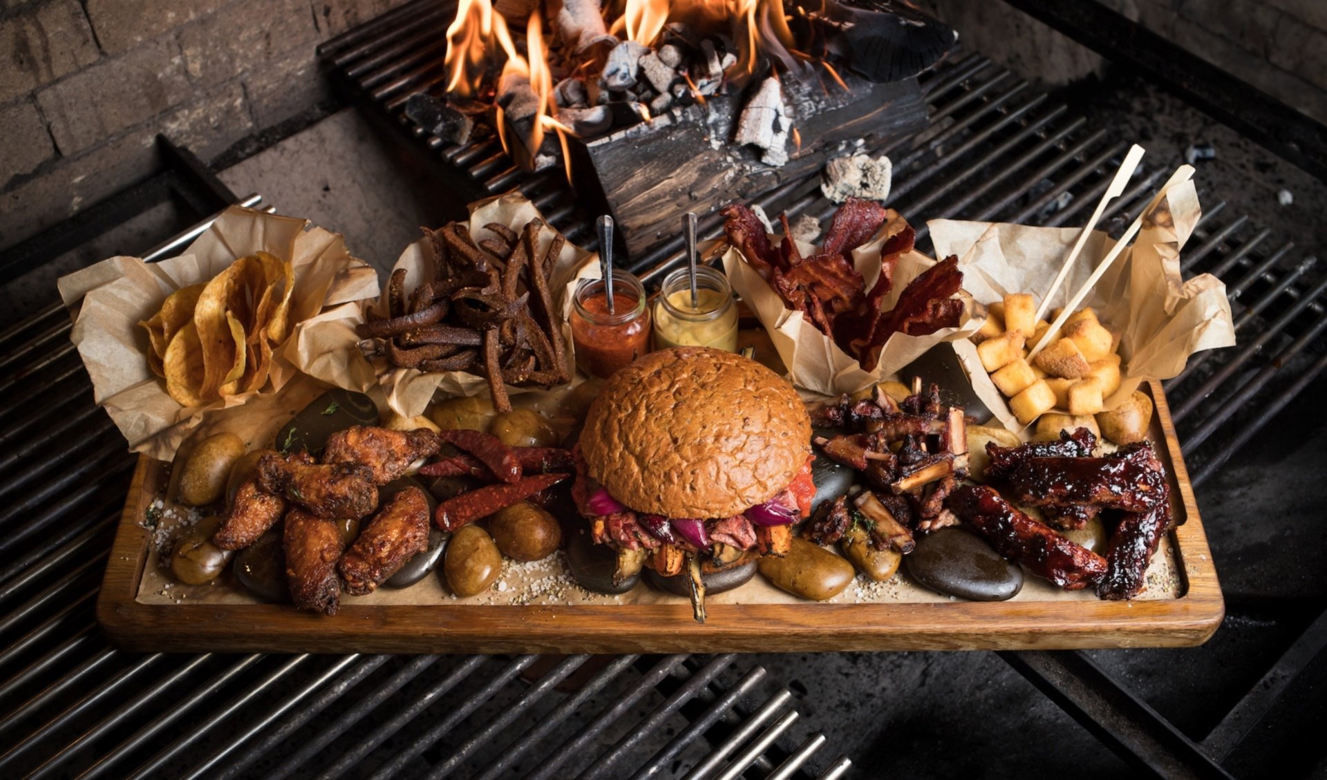 HD PC desktop wallpaper featuring a delicious, loaded burger centerpiece surrounded by wings, ribs, fries, and dipping sauces over a fiery grill background.