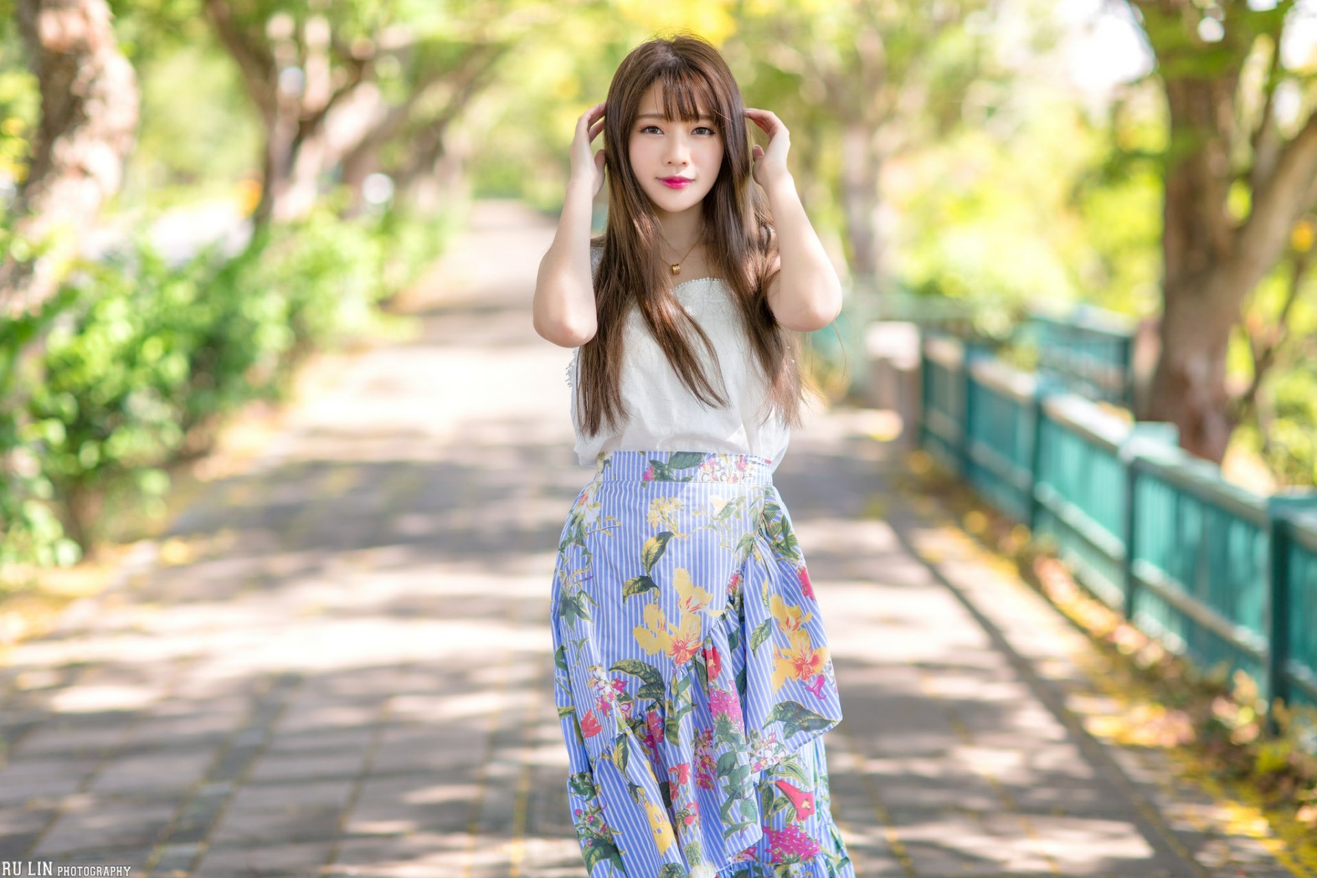 HD desktop wallpaper of an Asian woman with long brunette hair and lipstick, standing on a sunlit path with blurred greenery in the background, showcasing depth of field.