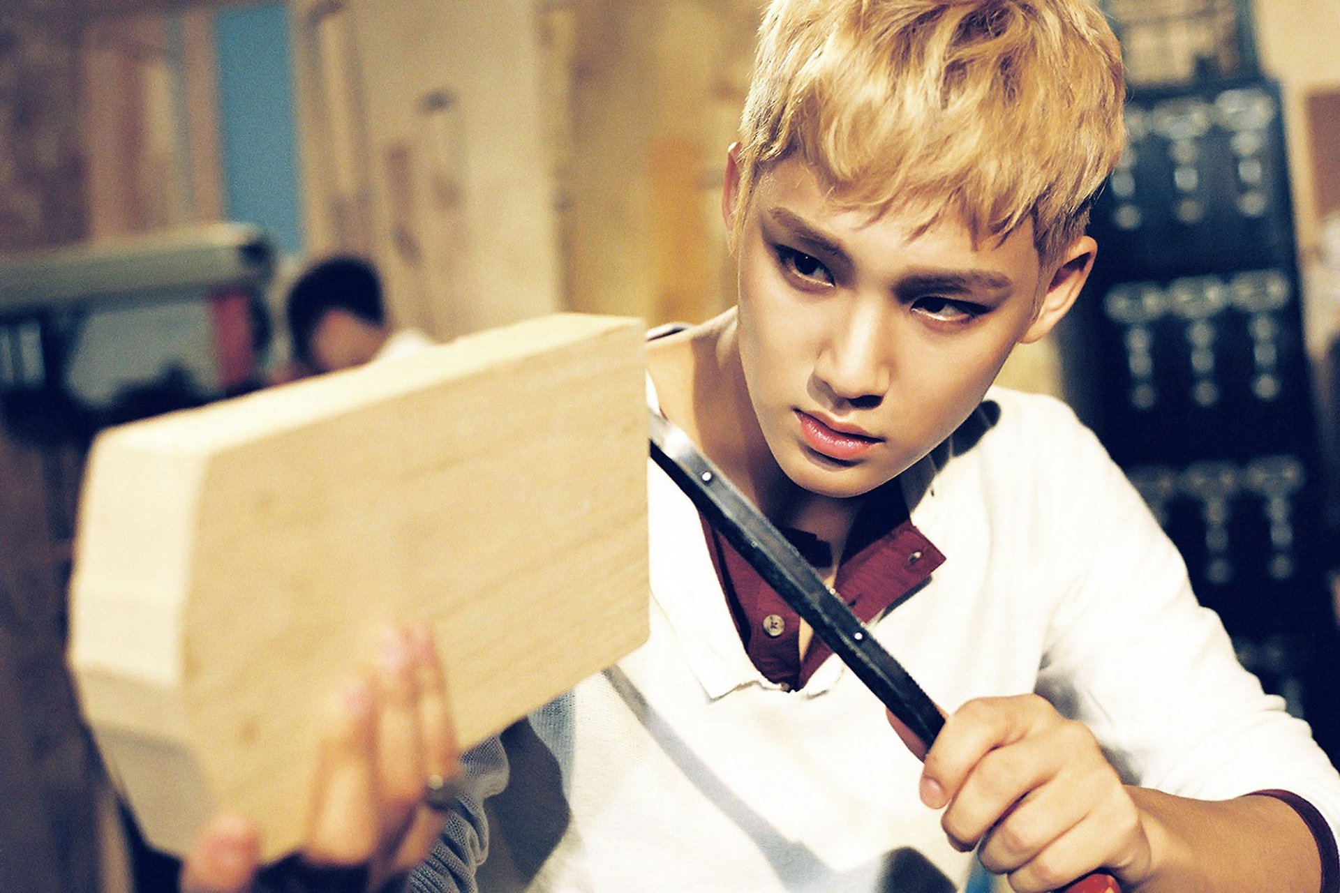 HD PC desktop wallpaper of a blond K-pop idol intensely carving a wooden block in a dim workshop, music-themed background.