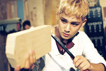 HD PC desktop wallpaper of a blond K-pop idol intensely carving a wooden block in a dim workshop, music-themed background.