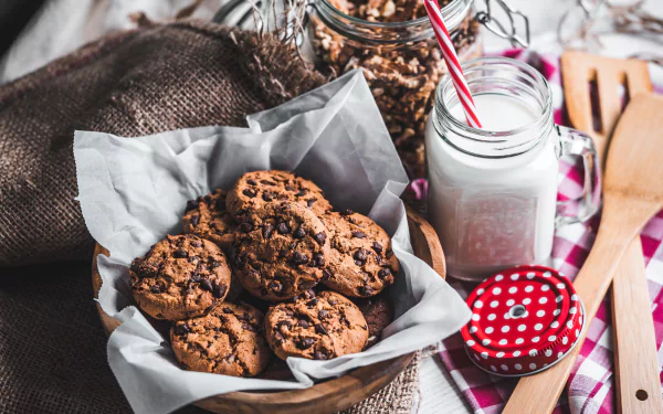 A still life 4K Ultra HD wallpaper featuring a basket of chocolate chip cookies alongside a jar of milk with a red-striped straw.